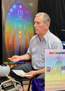 An older man sat down talking to someone out of shot with a banner behind him displaying a figure revealing the different points for each chakra.